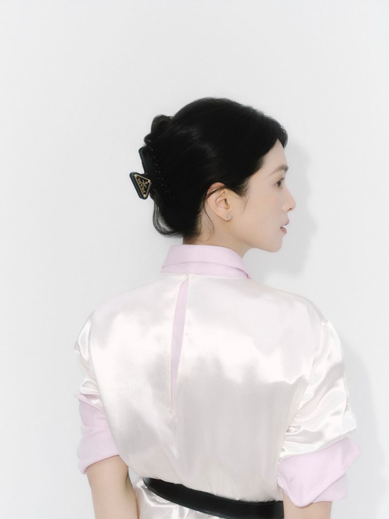 Brunette woman from behind wearing light pink and white shirt and black hair clip with logo against a white studio backdrop.