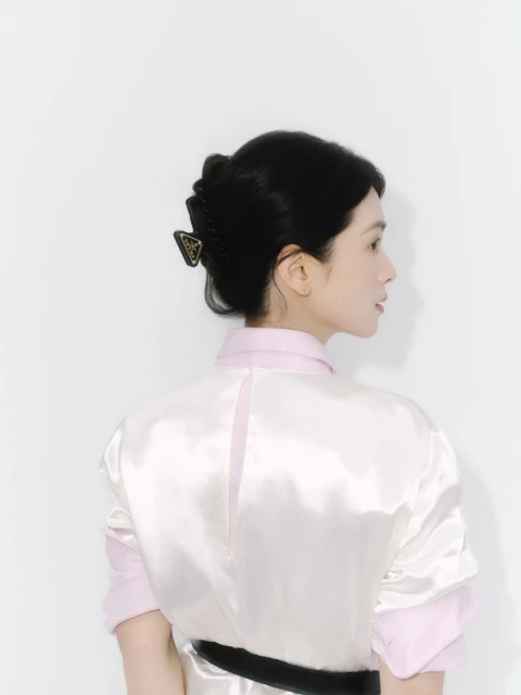 Brunette woman from behind wearing light pink and white shirt and black hair clip with logo against a white studio backdrop.