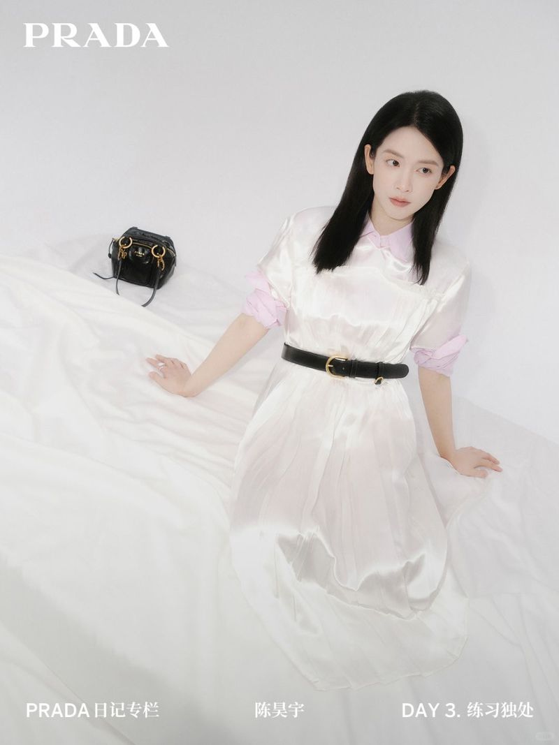 A woman in a white dress sits on white fabric next to a black purse and the Prada logo, in a fashion advertisement campaign.