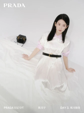 A woman in a white dress sits on white fabric next to a black purse and the Prada logo, in a fashion advertisement campaign.