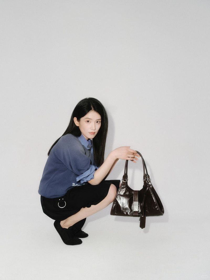 Young woman in blue shirt and black skirt crouches indoors, holding a dark brown leather handbag, captured in a medium shot.
