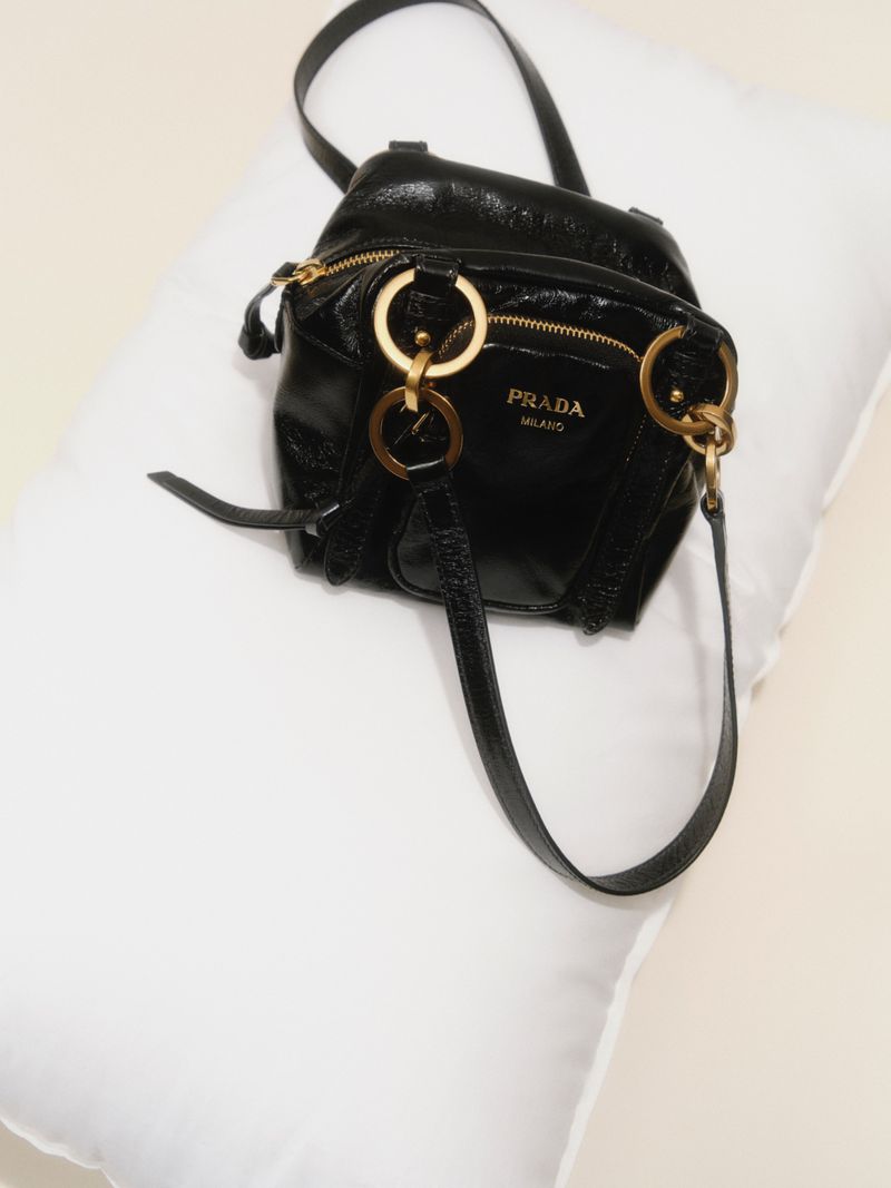 Elevated shot of a black Prada handbag featuring gold hardware and the Prada logo, set against a soft white pillow background.
