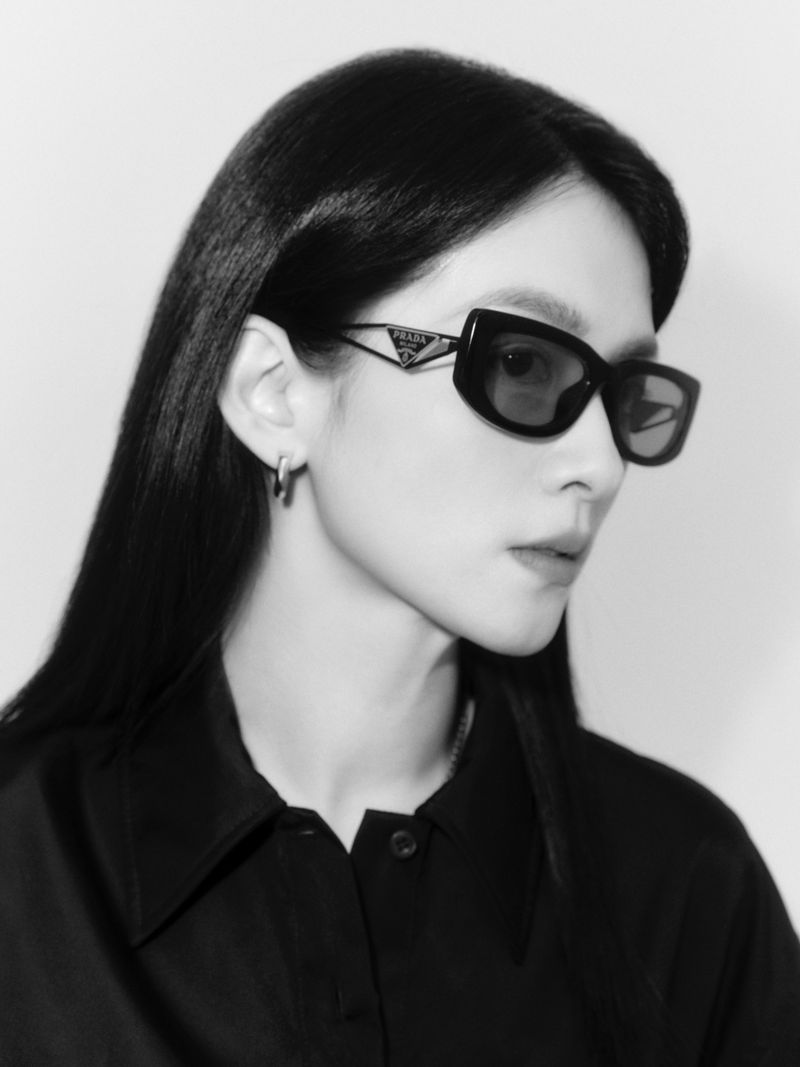 Portrait of a woman wearing dark Prada sunglasses and a black collared shirt against a plain background
