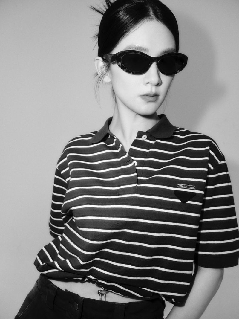 Black-and-white portrait of a young adult woman with sunglasses, wearing a horizontally striped polo shirt, standing in a studio.