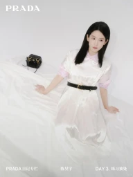 Woman with long dark hair wearing a white dress and black belt sitting on a white seamless backdrop with a black handbag