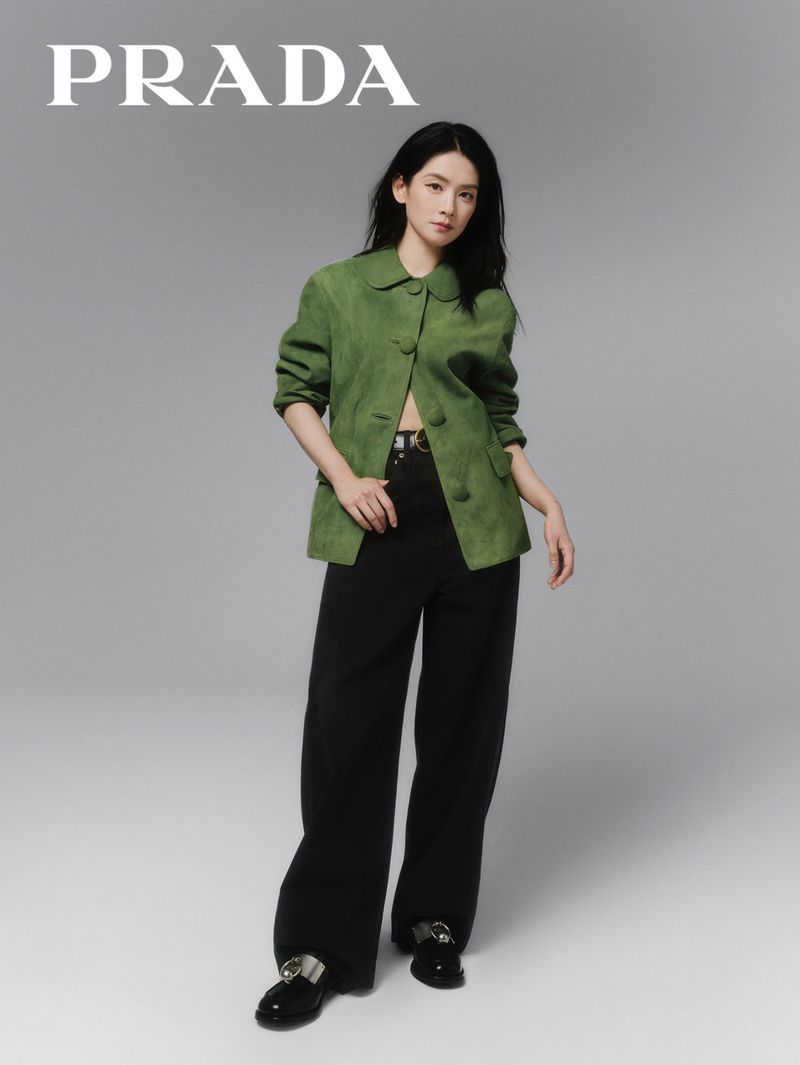Full-length portrait of a brunette adult female in a green blazer and black pants standing in a neutral grey photo studio setting.
