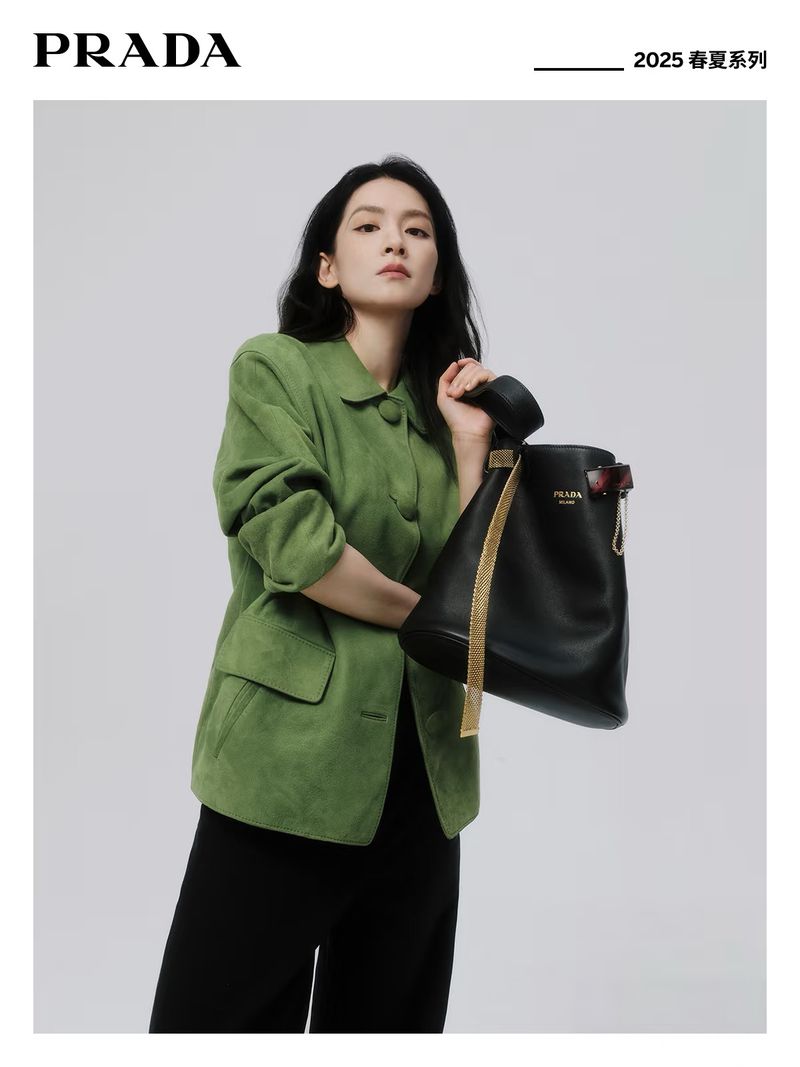A woman models a green jacket with a black leather bucket bag in a studio setting for a fashion advertisement, minimalistic background.