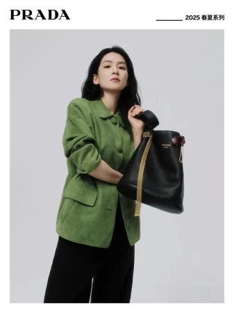 A woman models a green jacket with a black leather bucket bag in a studio setting for a fashion advertisement, minimalistic background.