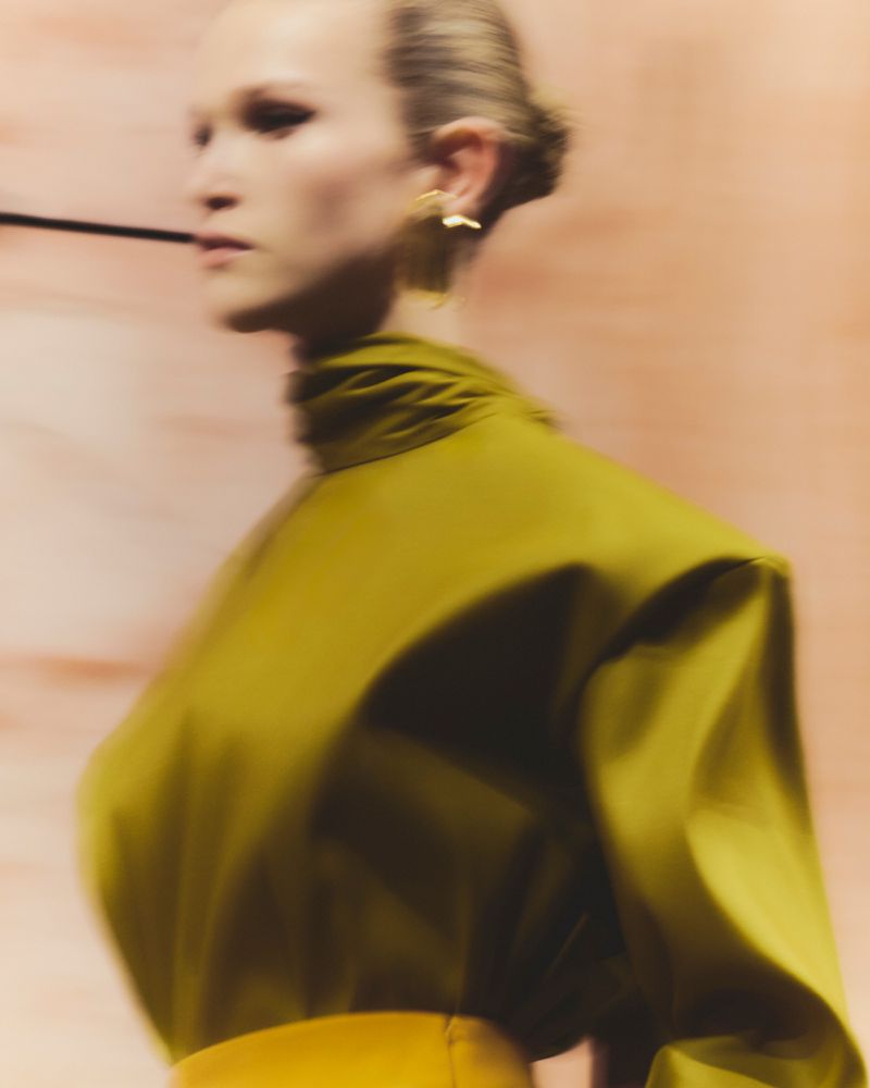 Woman in olive green high-neck blouse and yellow skirt in blurred close-up fashion shot, wearing earrings against a peach background.
