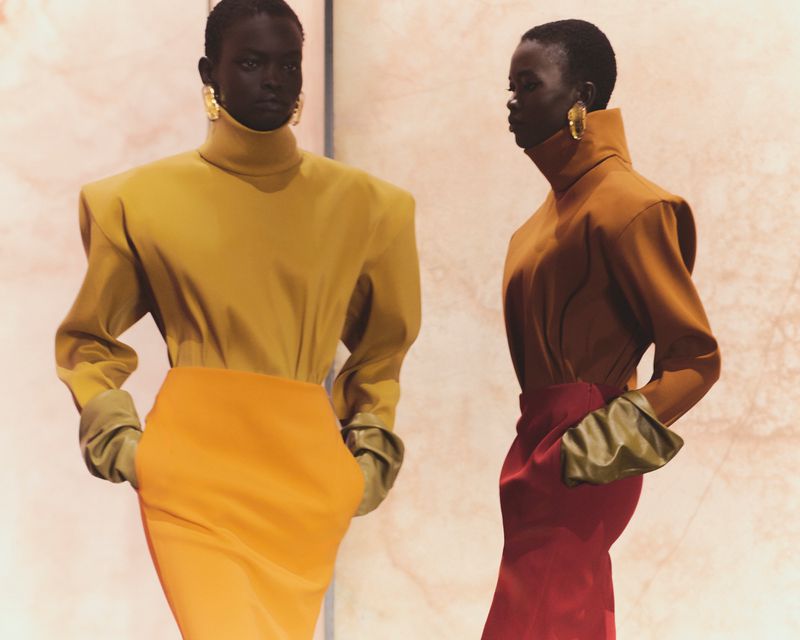 Two dark-skinned fashion models showcase stylish outfits with structured tops, pencil skirts, and bold gold earrings against a blurred backdrop.