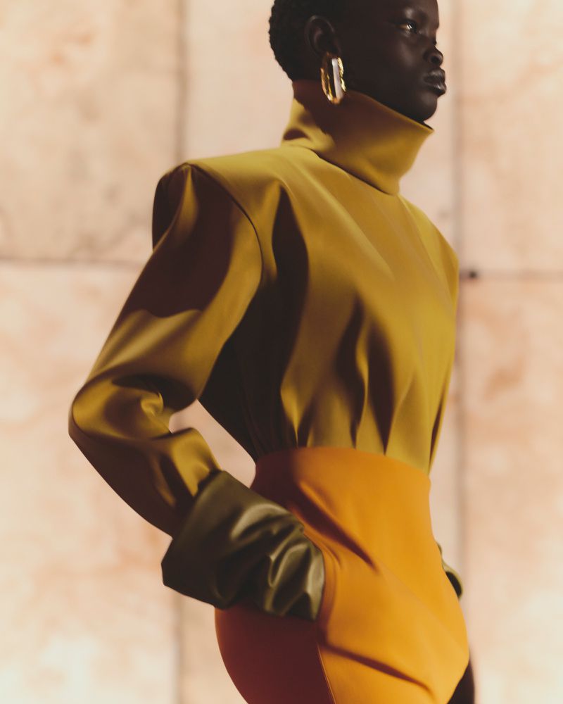 Fashion shot of an African American female model in a mustard turtleneck and orange skirt. Studio lighting and a peach-toned backdrop.