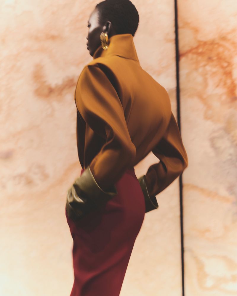 An adult female model posing, wearing a bronze top and dark red skirt, with a neutral backdrop, emphasizing fashion and portraiture.