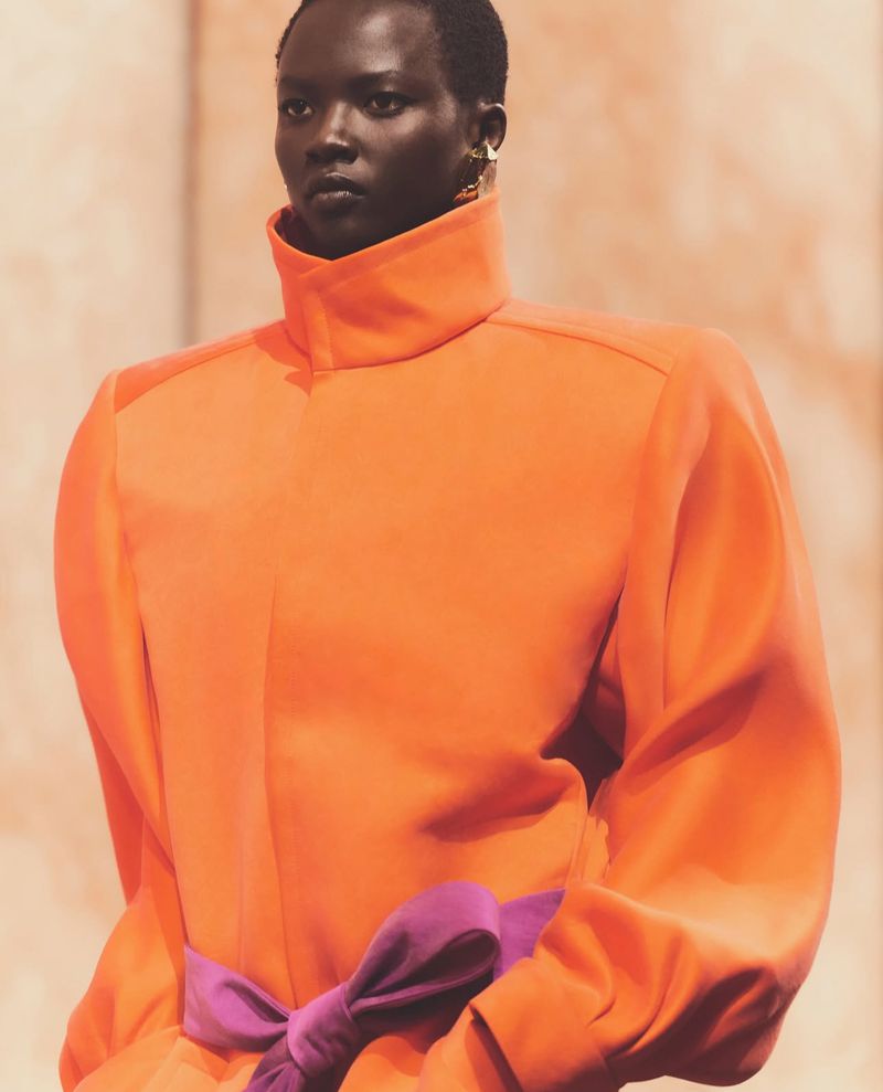 Close-up of a dark-skinned adult female fashion model in an orange high-neck dress with a purple bow, posing indoors against a light background.