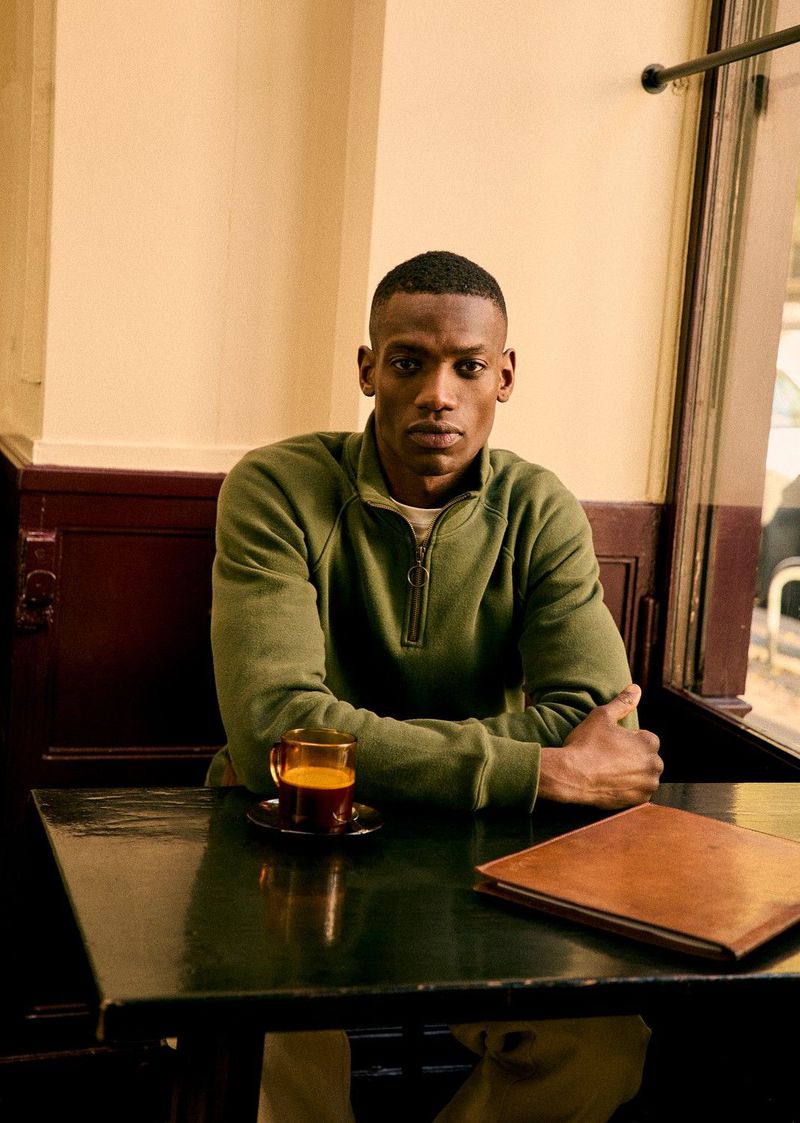 An adult male sits at a black table inside a cafe with a drink and book, wearing an olive green sweatshirt in a medium shot.