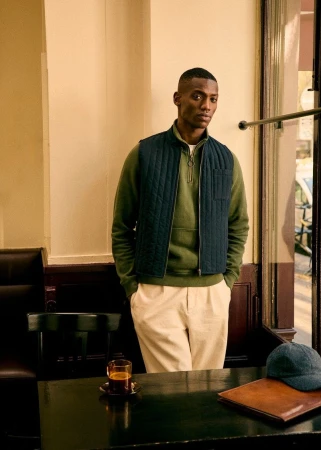 A young Black man wearing a quilted vest and olive sweatshirt sits at a wooden table next to a window inside a cafe.