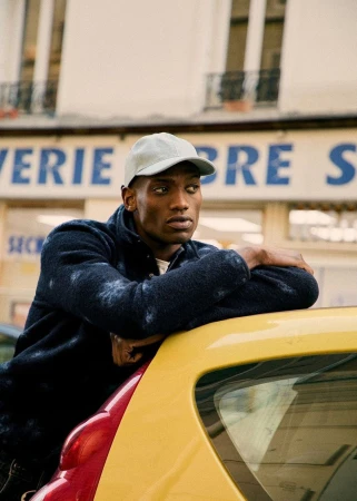 Black man wearing a grey cap and dark fleece leaning on a yellow car outside a building in Paris