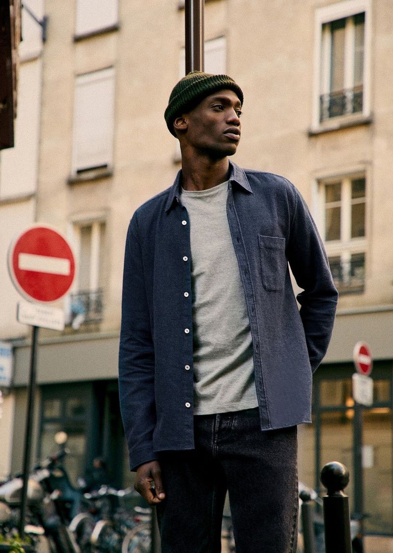 Man in casual fashion stands on a city street in shirt, t-shirt, dark jeans, and knit cap, with urban background and natural lighting.