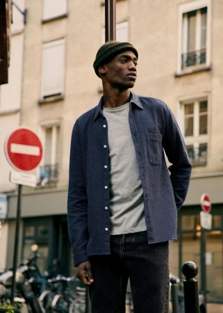 Man in casual fashion stands on a city street in shirt, t-shirt, dark jeans, and knit cap, with urban background and natural lighting.