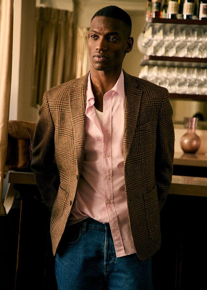 Portrait of an African American male wearing a brown plaid blazer, pink dress shirt, and denim jeans standing indoors with neutral background.
