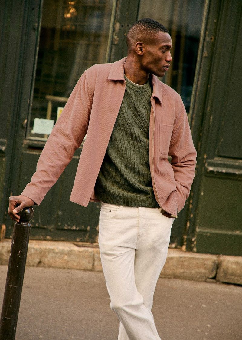 Man in a pink jacket and white jeans stands near a building, wearing a green shirt, posing casually against an urban backdrop.