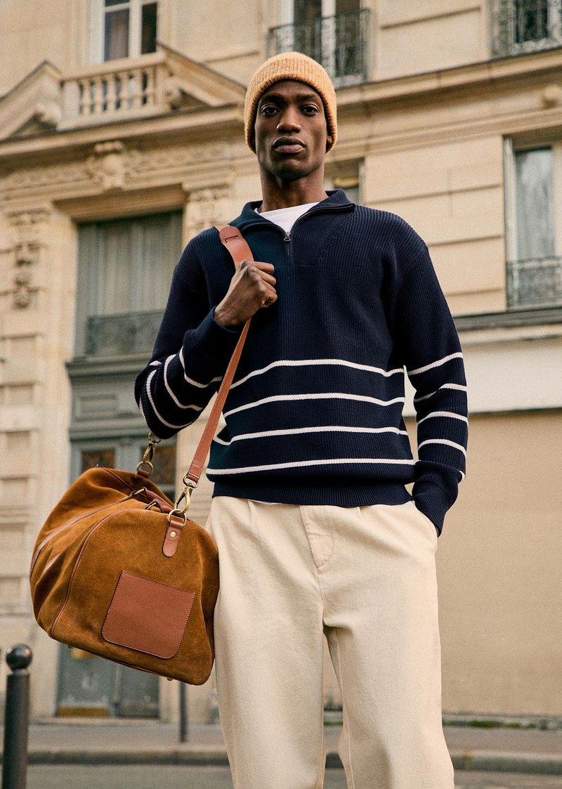 Adult male in navy striped sweater carries tan duffel bag in urban setting near building with architectural detail on sunny day.