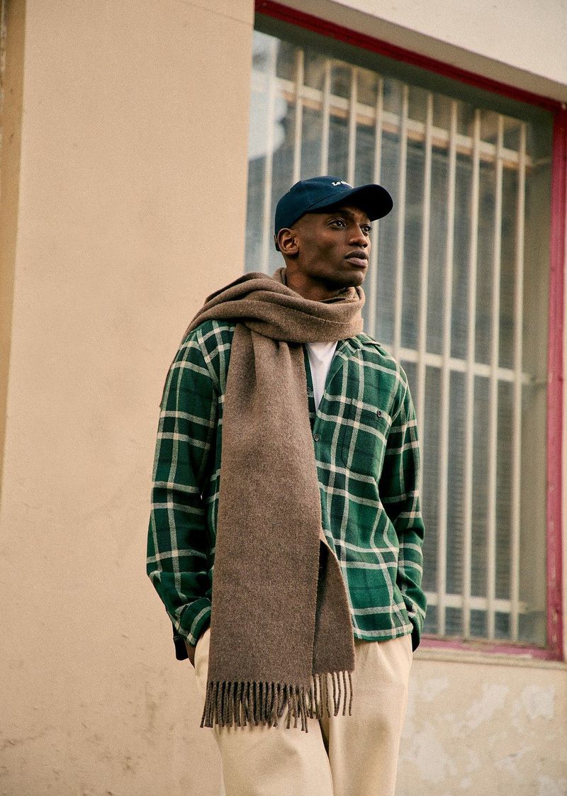Portrait of a Black male model in a plaid shirt, scarf, and baseball cap, standing outdoors against a neutral wall with a window.