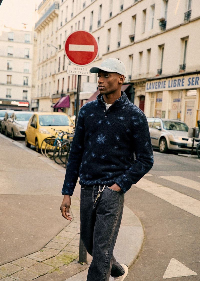 An adult man in a patterned top and jeans stands on a city sidewalk next to parked cars and buildings in an outdoor urban setting.