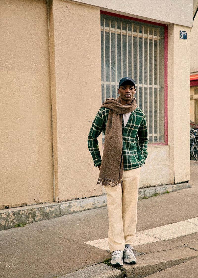 A young man stands on a city sidewalk wearing a green plaid cardigan, cream-colored pants, sneakers, scarf, and baseball cap.