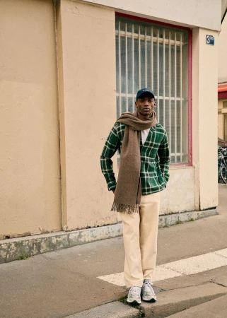 A young man stands on a city sidewalk wearing a green plaid cardigan, cream-colored pants, sneakers, scarf, and baseball cap.
