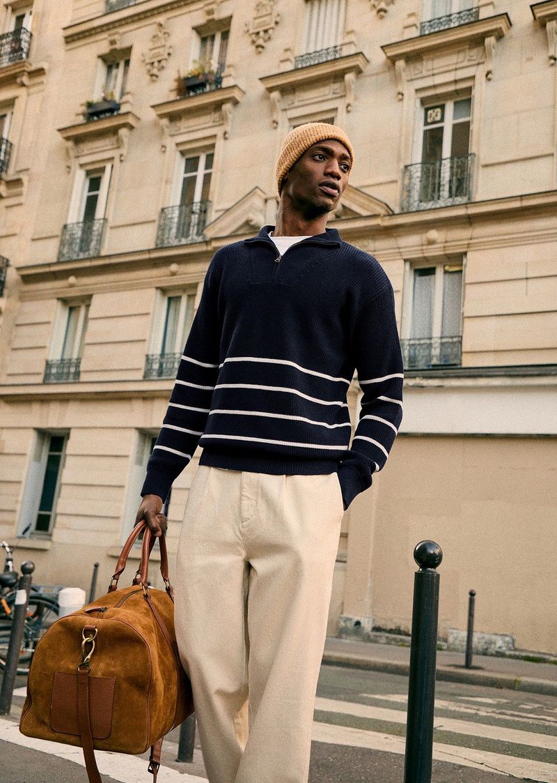 Man in navy striped sweater walking on urban street with brown duffel bag. He is wearing tan pants and beanie in city environment.