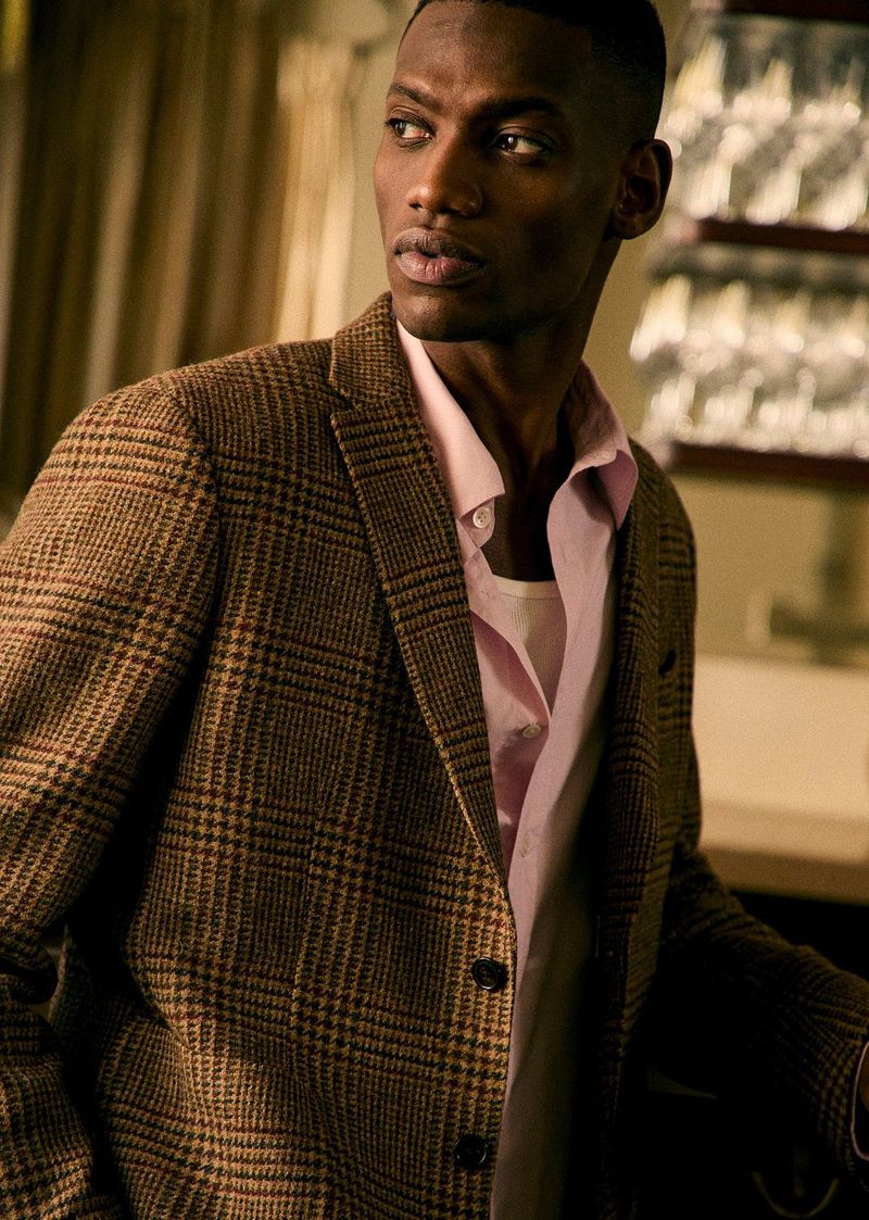 An adult male wearing a brown plaid blazer over a pink button-down shirt poses indoors with an out-of-focus background.
