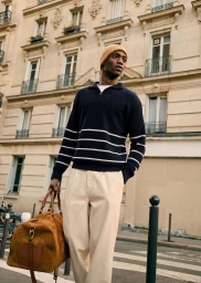 Model wearing a navy striped sweater, beige trousers, and a yellow beanie, holding a brown duffel bag on a city street.