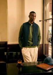Black man standing in a café wearing a green sweater and navy quilted vest for Octobre Editions Feb 2025.