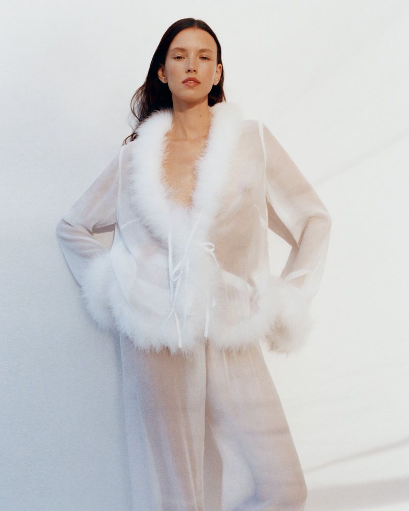 Woman in sheer white robe with faux fur trim poses against white wall in studio lighting. Fashion, beauty, and style shoot.