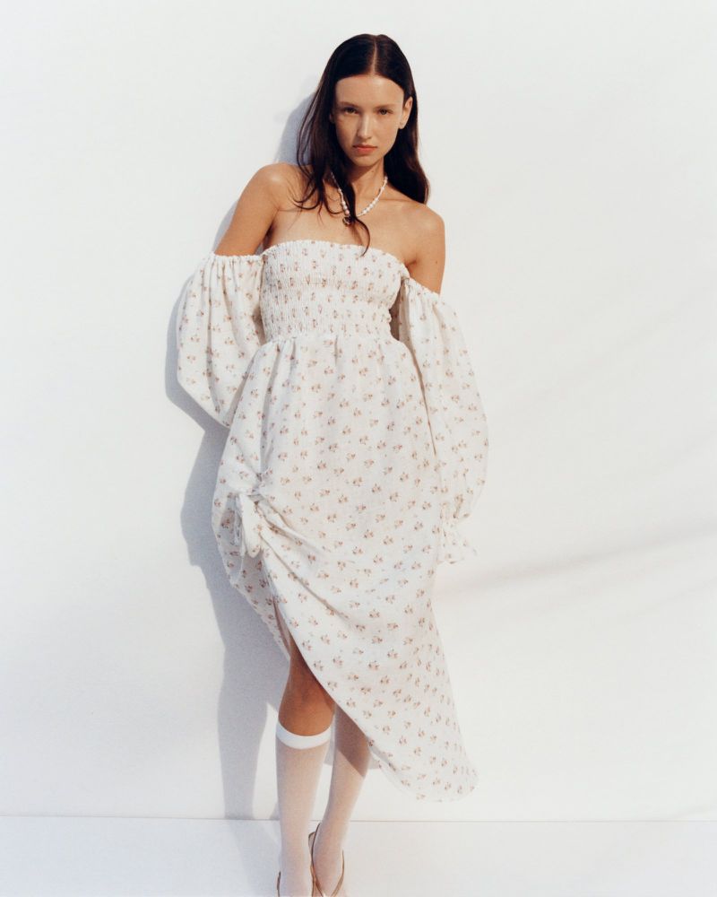 A woman in a white floral dress stands against a white wall, creating a clean and fashionable full shot in a studio setting.