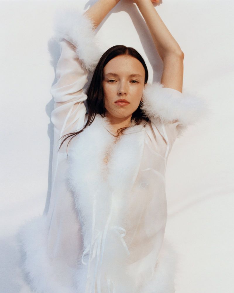 Brunette woman in sheer white robe with feathered trim, arms overhead, in a fashion portrait against a white studio backdrop.