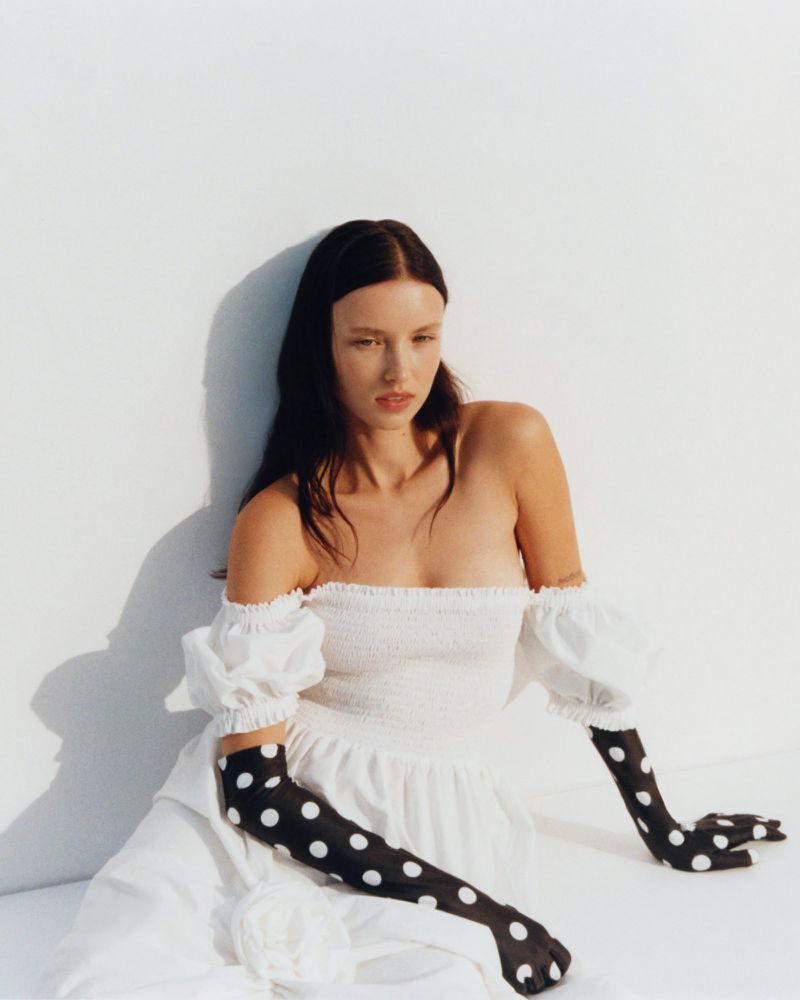 Woman poses in a white off-shoulder dress and long black polka dot gloves against a white background in a fashion photography studio shot.