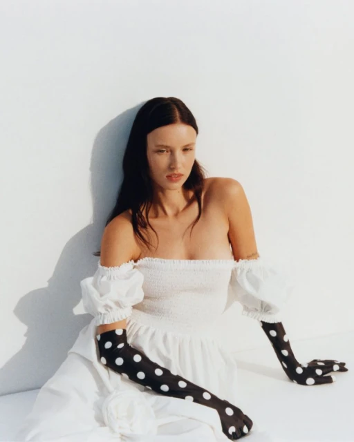 Woman poses in a white off-shoulder dress and long black polka dot gloves against a white background in a fashion photography studio shot.