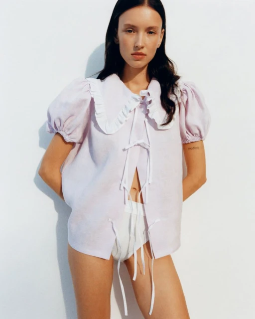 A young woman poses in a lilac top with a frilly white collar, accessorized with string ties, against a plain white background.