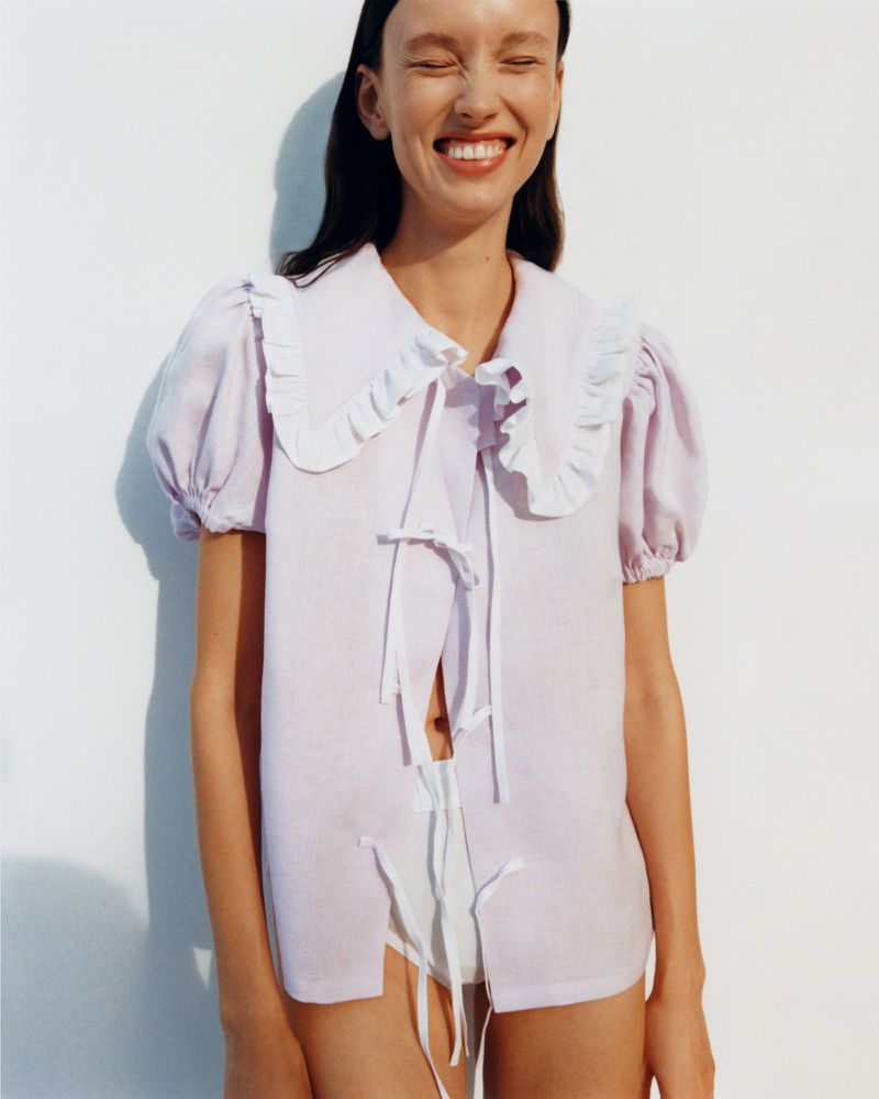 Smiling woman in a lavender ruffle-collared blouse with white tie closures, posing in front of a white background in a studio shot.