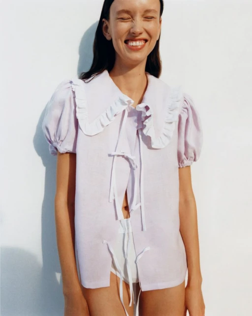 Smiling woman in a lavender ruffle-collared blouse with white tie closures, posing in front of a white background in a studio shot.