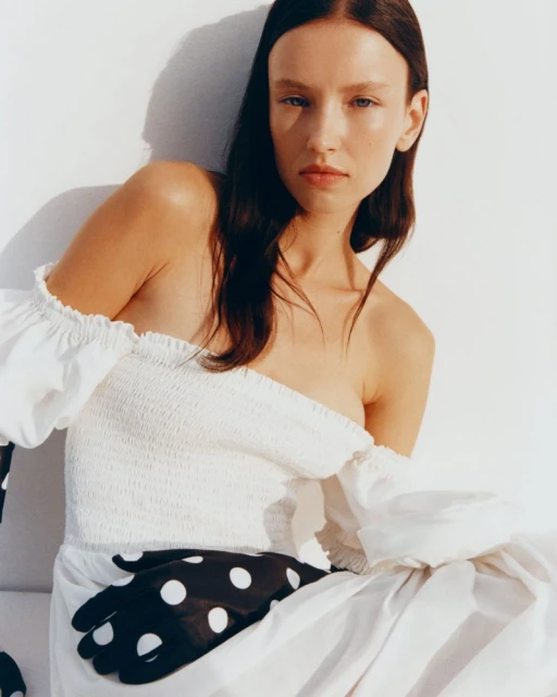 A model with brunette hair poses in a white dress with black and white polka dot gloves against a bright white background in a studio.
