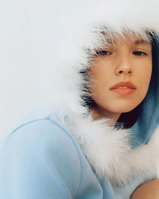 Close-up of a young woman with light skin wearing a light blue hoodie with a white fur trim around the hood, neutral expression, and white background.