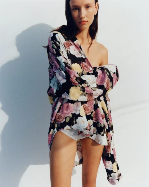 Young woman poses in floral print shirt against a white wall, displaying a relaxed and fashionable style, captured in a medium shot.