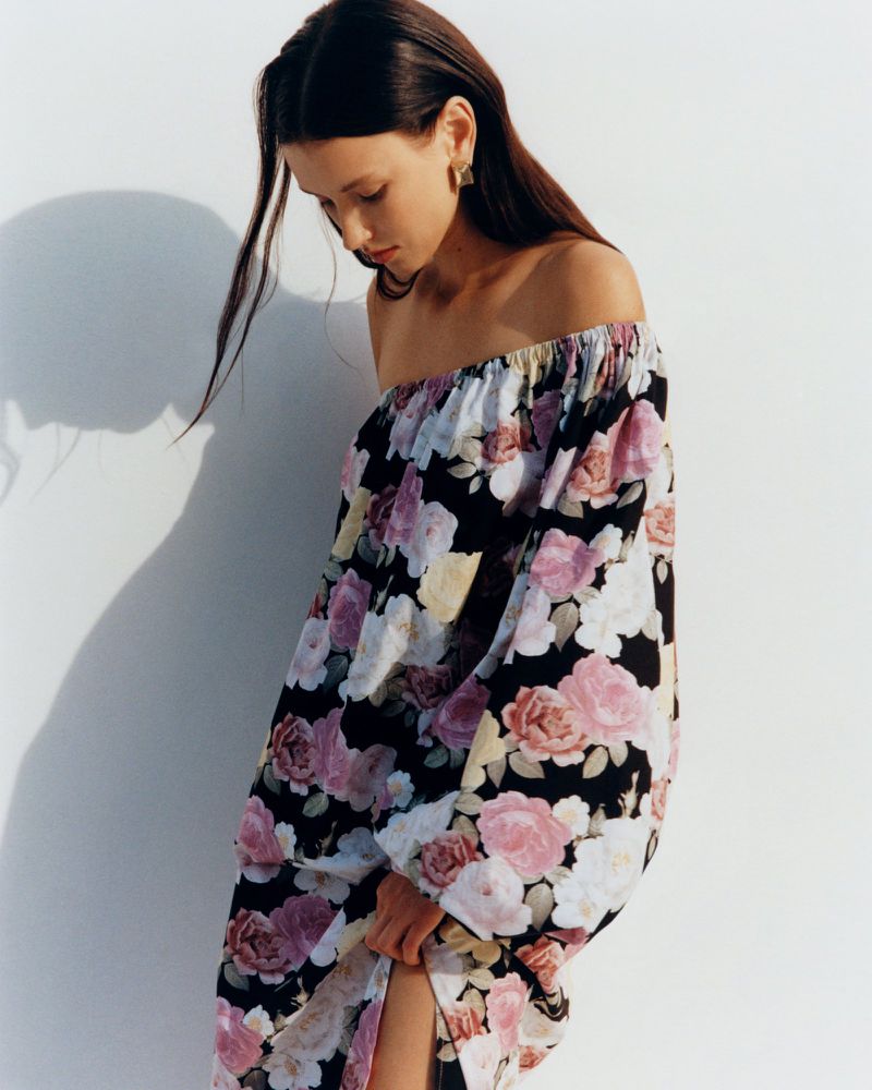 A young woman poses in a black, pink, and white floral off-the-shoulder dress against a white wall, looking down with soft, natural lighting.