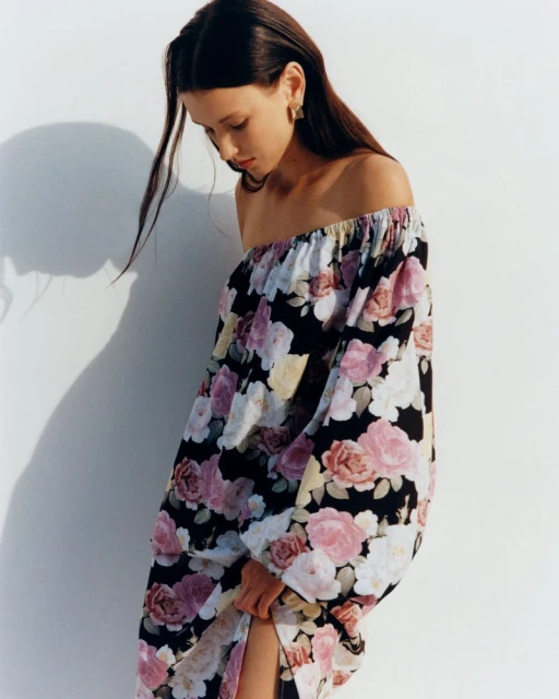 A young woman poses in a black, pink, and white floral off-the-shoulder dress against a white wall, looking down with soft, natural lighting.