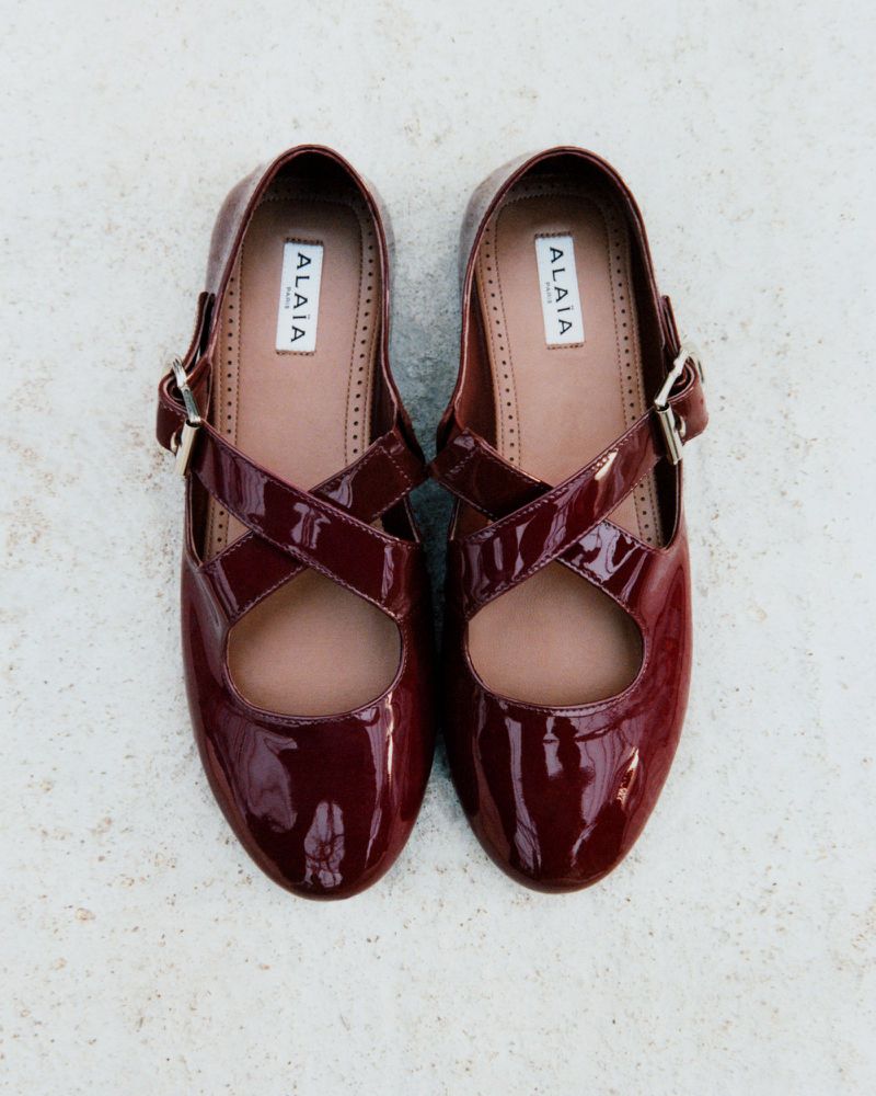 Burgundy Alaïa Mary Jane ballet flats featuring glossy patent leather, cross-over straps, and a buckle, set against a textured background.