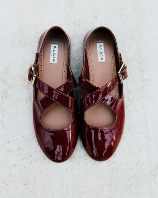 Burgundy Alaïa Mary Jane ballet flats featuring glossy patent leather, cross-over straps, and a buckle, set against a textured background.