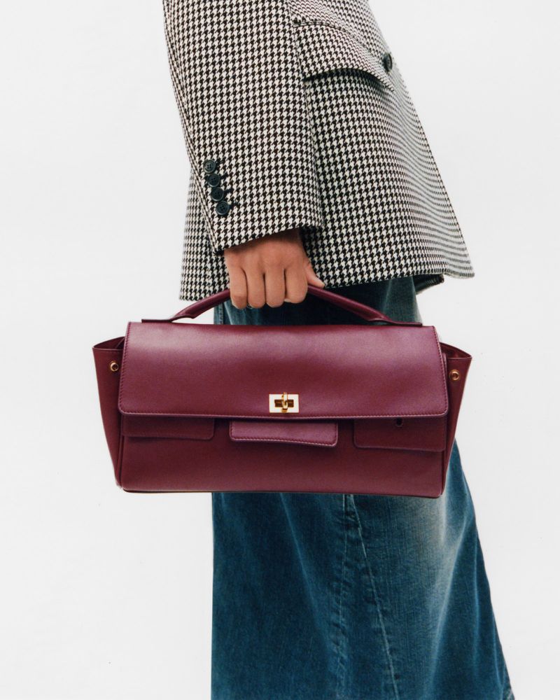 Burgundy leather satchel bag held by a person wearing a houndstooth blazer and denim skirt, set against a plain white backdrop.