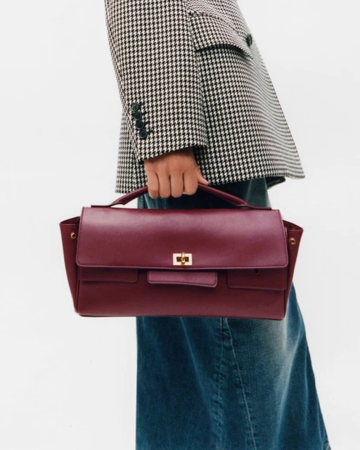 Burgundy leather satchel bag held by a person wearing a houndstooth blazer and denim skirt, set against a plain white backdrop.